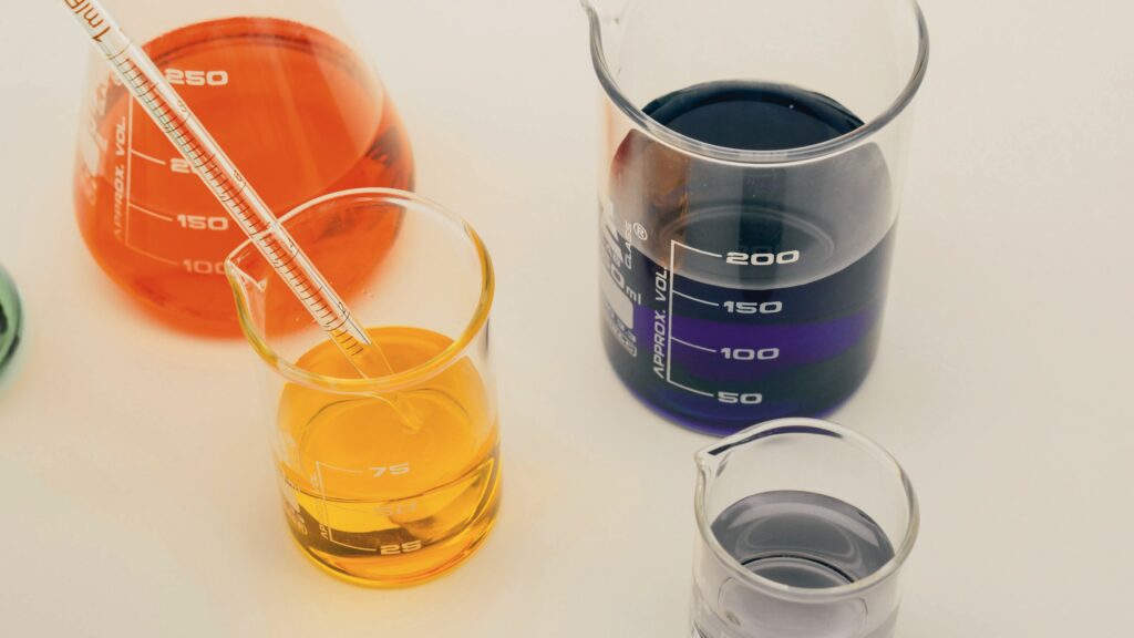 Laboratory beakers with orange, yellow, and purple liquids representing the range of peptides — including BPC-157, Ipamorelin, and Sermorelin — used in peptide therapy in Cumming, GA at Good Chemistry.